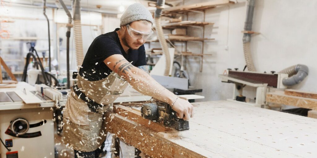 Carpenter working with electric planer on wooden plank in workshop.