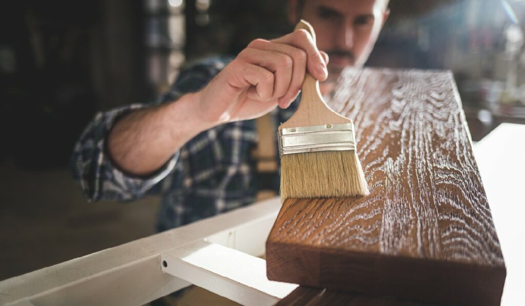 Close up of paintbrush applies paint or varnish on wooden board in carpentry workshop