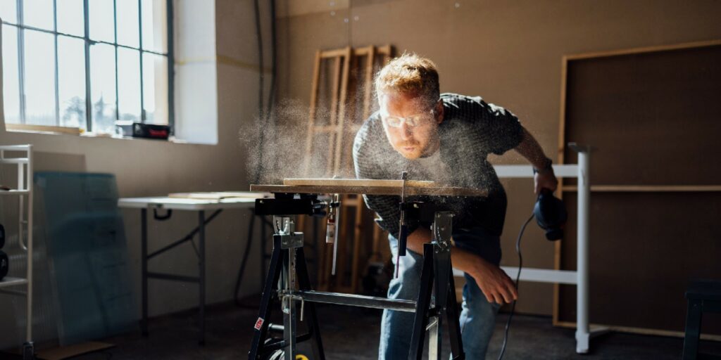 Carpenter Blowing Dust From Wooden Plank