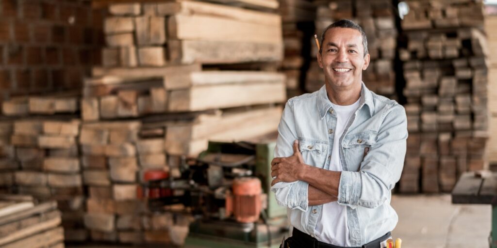 Friendly carpenter at his workshop with piles of wood looking very happy