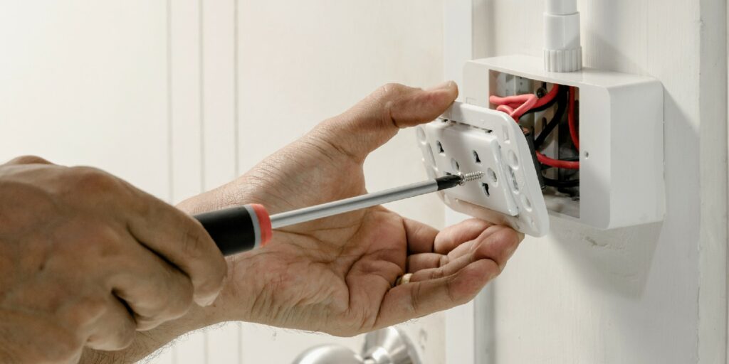 The electrician is using a screwdriver to attach the power cord to the wall outlet.