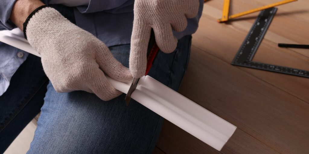 Man Cutting Foam Crown Molding with Utility Knife near Wooden Ta